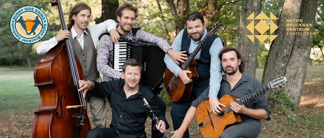 Fünf Musiker posieren für ein Gruppenfoto in einem Wald, jeder spielt ein Musikinstrument. Ein Mann mit Bart hält eine Gitarre, ein anderer spielt Akkordeon, während die anderen Klarinette, Gitarre und Trompete spielen.