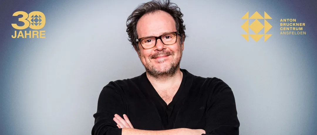 Porträt eines Mannes mit lockigem Haar und Bart, der eine Brille und ein schwarzes Hemd trägt. Er lächelt mit verschränkten Armen. Der Hintergrund ist grau.