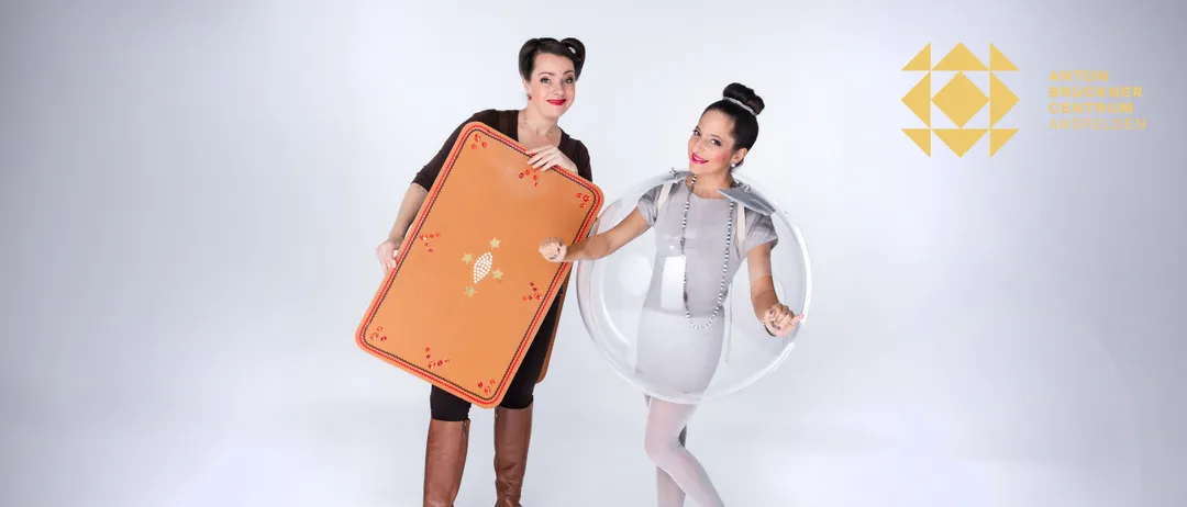 Zwei Frauen stehen nebeneinander. Eine hält eine große, bunte Matte mit einem Sternmuster, während die andere ein durchsichtiges, blasenartiges Kleid trägt.