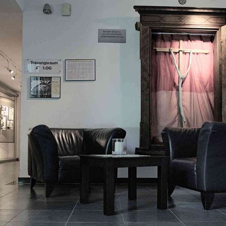 Ein Raum mit schwarzen Ledersofas, einem Holztisch, einem Kreuz und einem Holzrahmen mit einem Schild, das 'Traurungsraum 1.OG' auf der Wand steht.