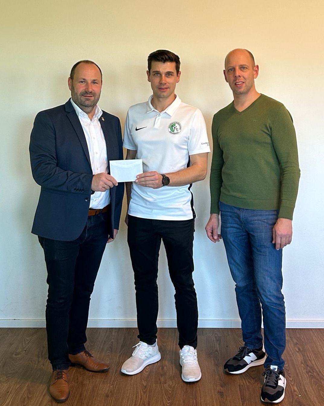 Drei Männer stehen nebeneinander vor einer weißen Wand, einer hält einen weißen Umschlag. Einer trägt einen Marineblazer, einer ein grünes V-Neck und der mittlere ein weißes Nike-Polo.