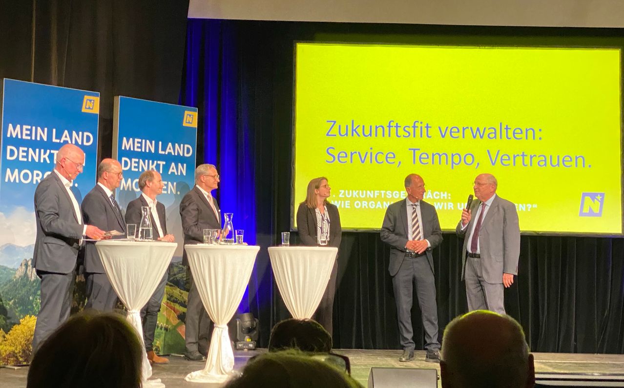 Fünf Personen stehen auf der Bühne, möglicherweise für eine Präsentation. Drei Männer und zwei Frauen sind in formeller Kleidung. Ein gelber Bildschirm mit Text steht hinter ihnen.