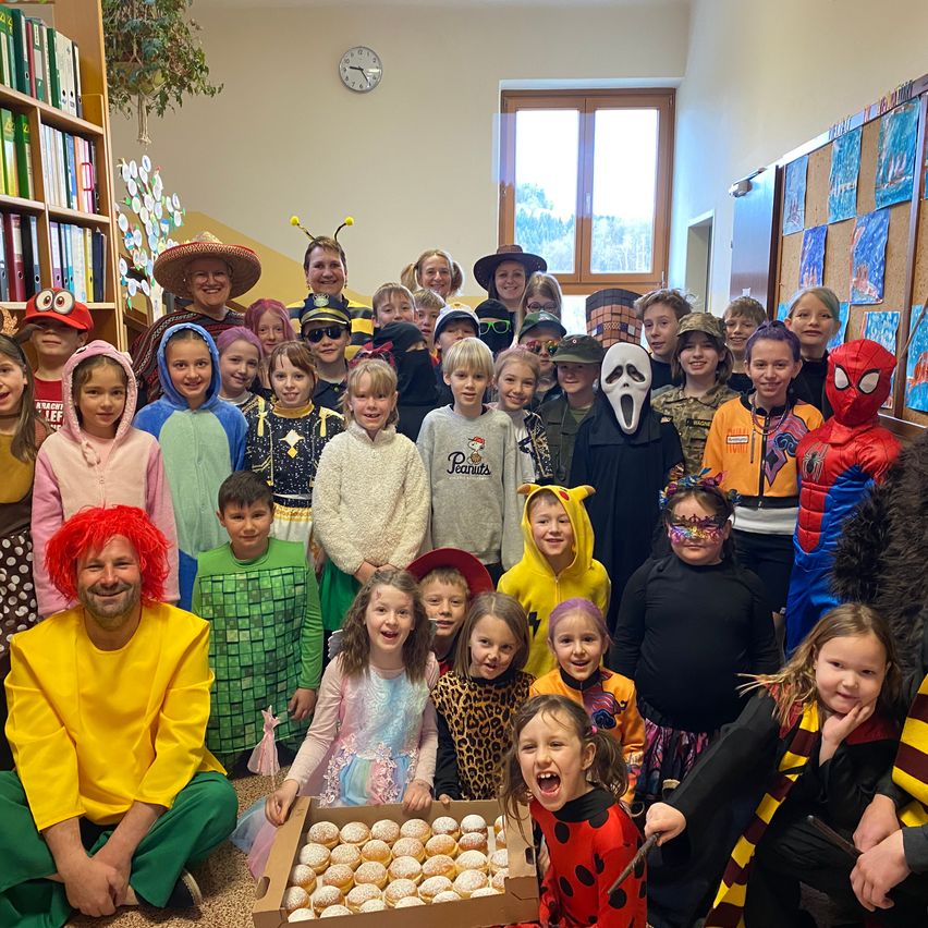 A group of children in various costumes, including Spiderman, a clown, and a ladybug, pose for a photo in a room with bookshelves, a clock, and a window. They are holding a box of doughnuts.