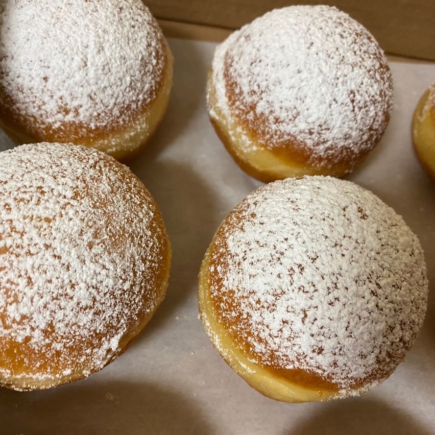 Five donuts with powdered sugar on top are in a box. Each donut is round and golden brown.
