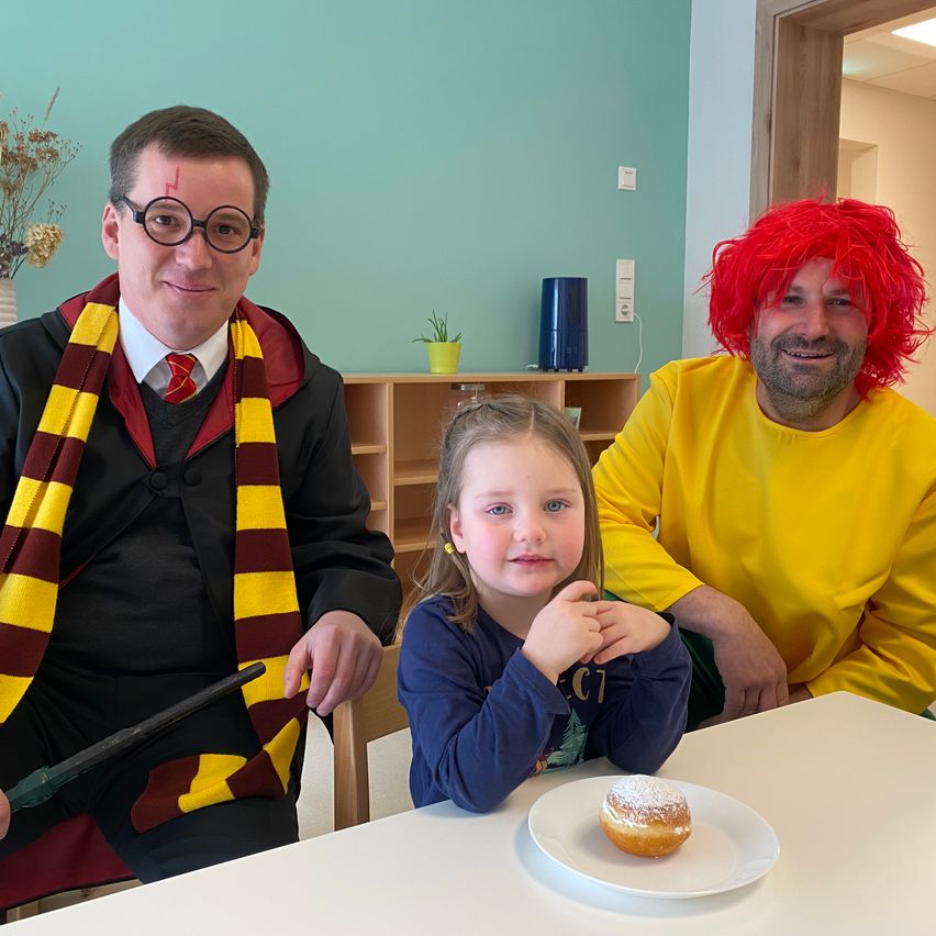 Three people dressed in costumes sit around a table with a child, a small potted plant, and a blue water jug in the background. One person holds a wand.