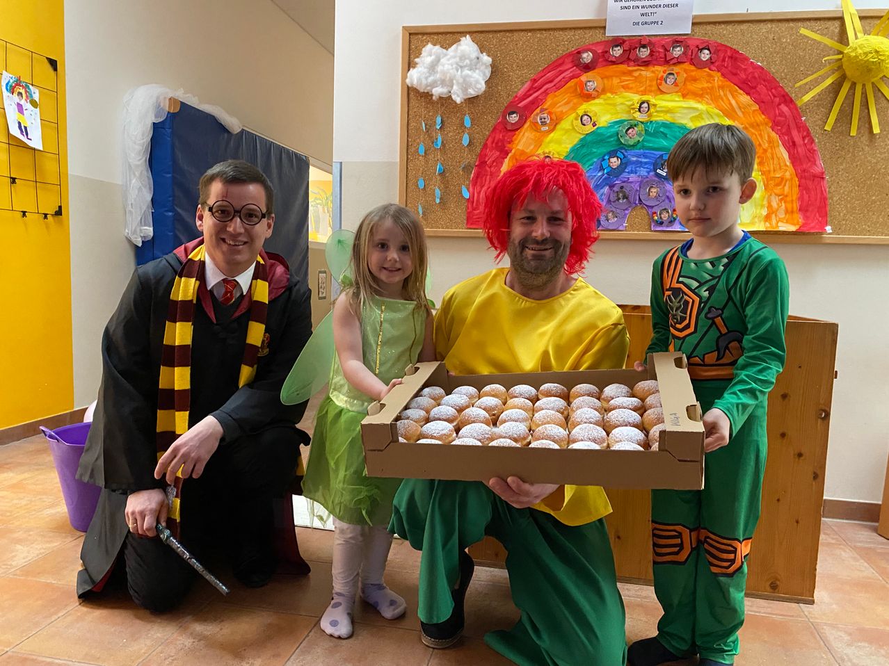 Four people dressed in costumes pose with a box of donuts. A boy holds a wand. A rainbow and cloud bulletin board is behind them.