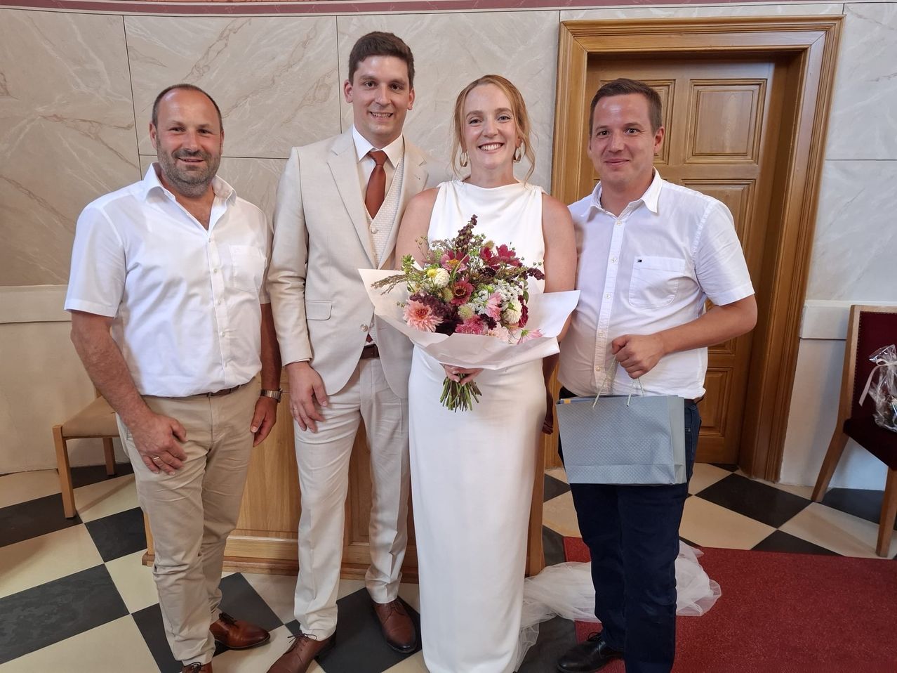 A woman in a white dress holding a bouquet of flowers stands between two men, one holding a paper bag and the other wearing a tie. They are all smiling and appear to be at a wedding.