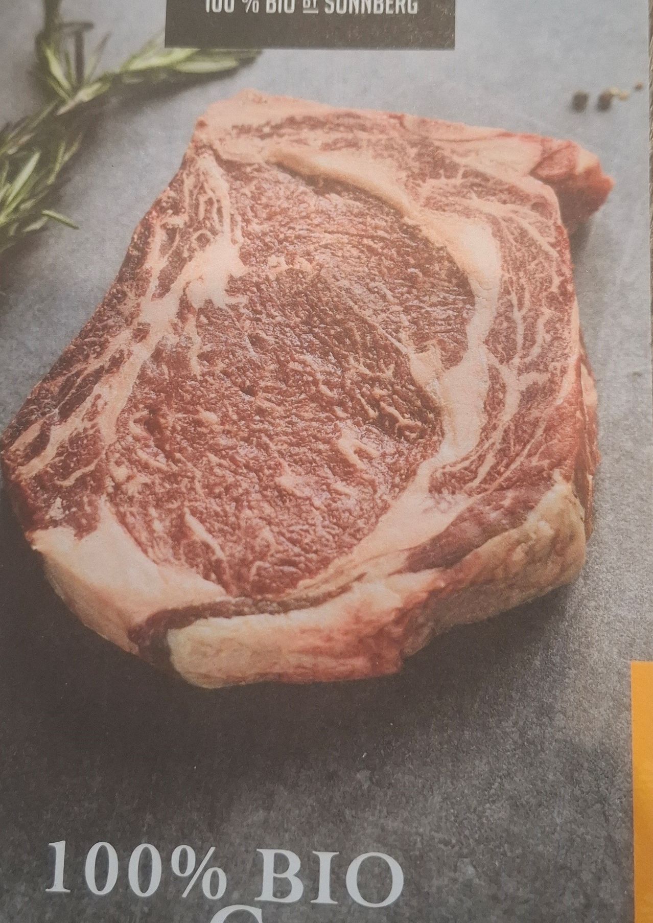 Ein roher Ribeye-Steak mit sichtbarer Marmorierung liegt auf einer grauen Oberfläche. Ein Rosmarinzweig ist links platziert.