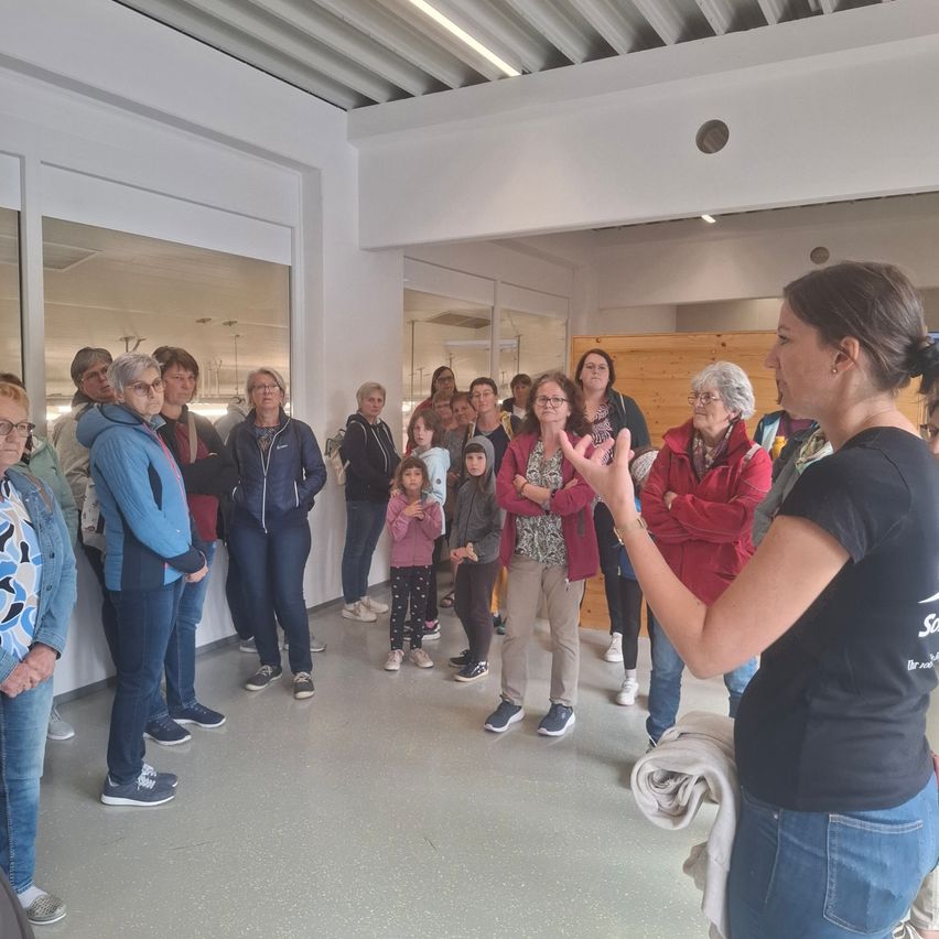 Eine Gruppe von Frauen und einigen Kindern versammelt sich in einem Raum. Eine Frau vor ihnen spricht zu ihnen. Der Raum hat weiße Wände, eine weiße Decke und Spiegel.