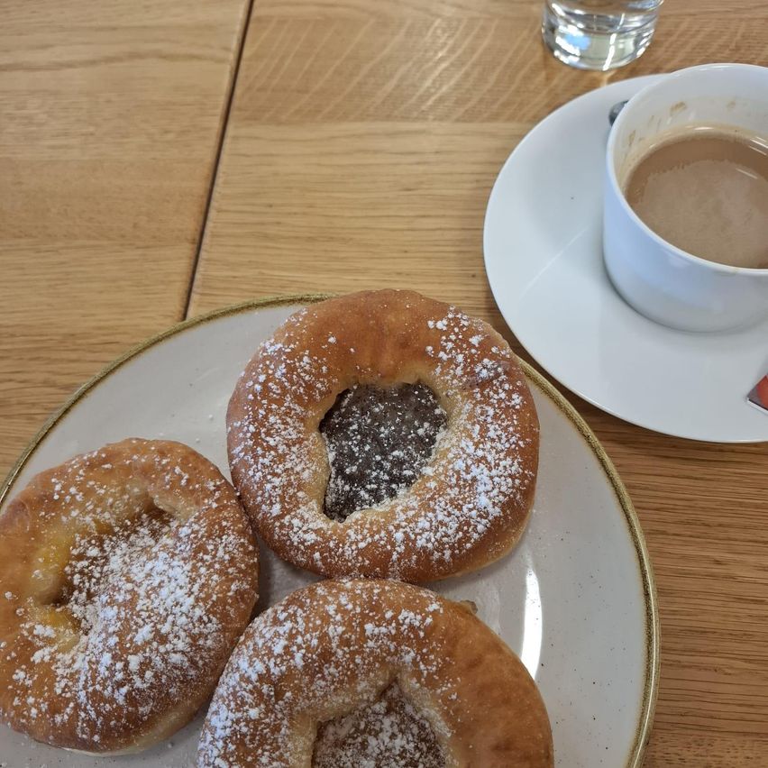 Ein Teller mit drei Donuts und einer Tasse Kaffee auf einer Untertasse. Die Donuts sind mit Puderzucker bestreut und haben Füllungen. Ein Glas Wasser steht auf dem Tisch.
