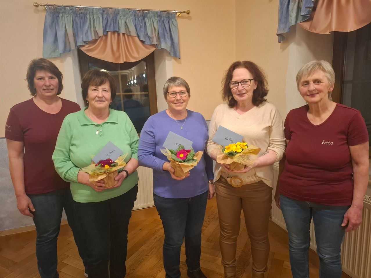 Fünf Frauen stehen lächelnd in einem Raum zusammen und halten Blumensträuße. Sie sind in verschiedenen Farben gekleidet. Hinter ihnen ist ein Fenster mit Vorhängen zu sehen.