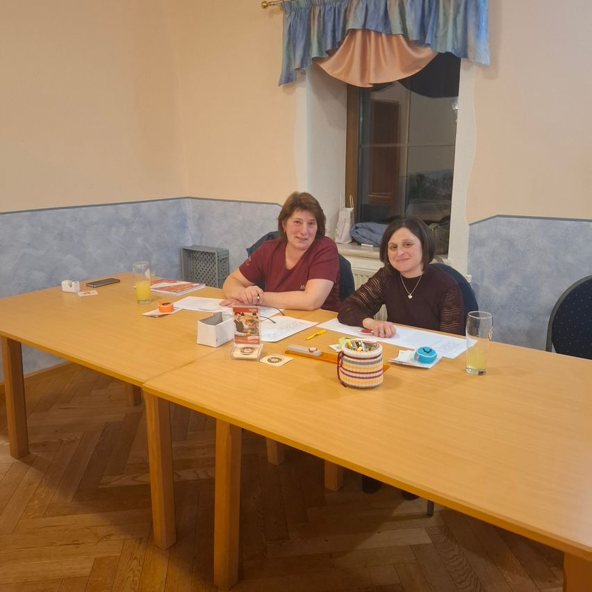 Zwei Frauen sitzen an einem Tisch mit Papieren, Gläsern mit Saft und einer Korb mit Gegenständen auf einem Schreibtisch. Sie lächeln und schauen in die Kamera.