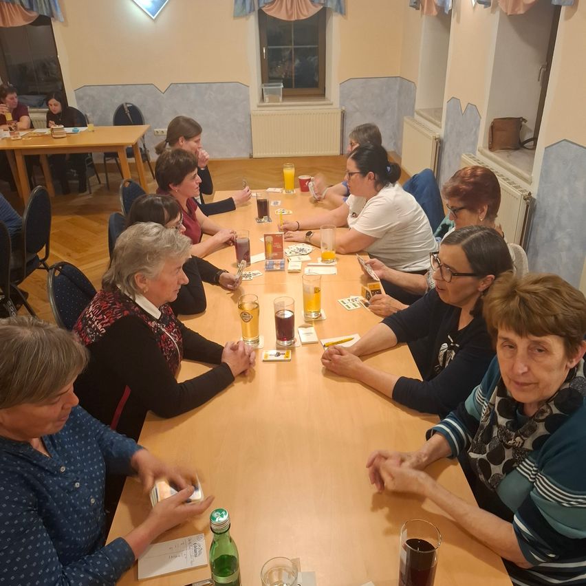 Eine Gruppe von Frauen sitzt um einen Tisch herum, möglicherweise spielt sie ein Kartenspiel. Der Tisch ist mit Karten, Gläsern und anderen Gegenständen übersät. Der Raum hat eine warme Atmosphäre mit Holzböden und weißen Wänden.