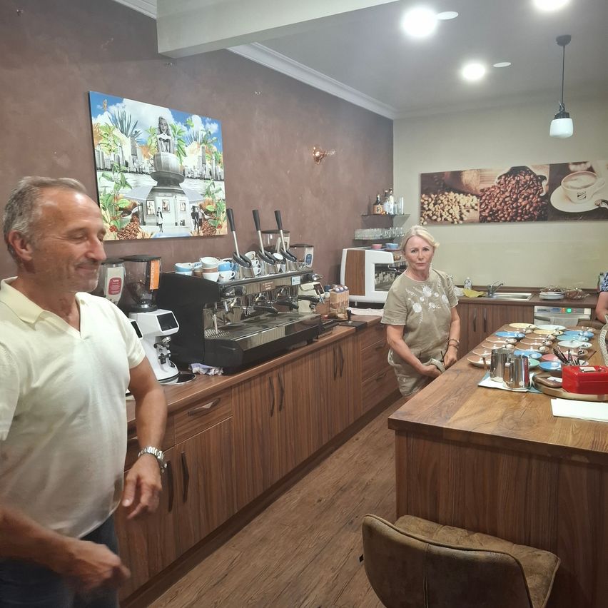 A man and a woman are standing in a coffee shop, with a coffee machine and various items on the counter.