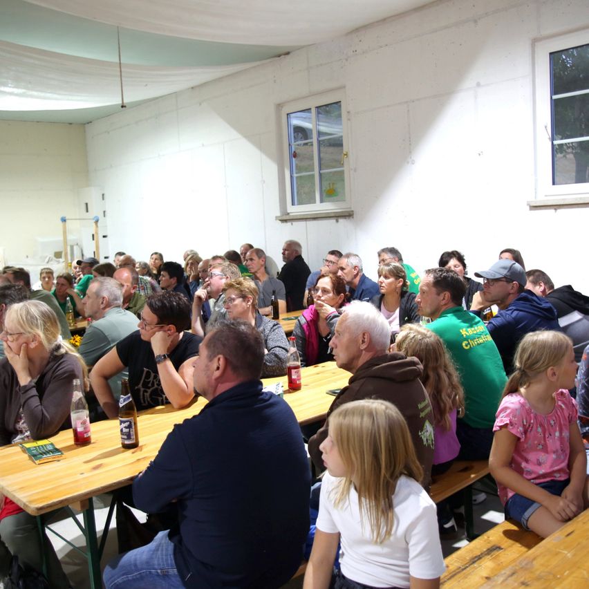 Eine große Gruppe von Menschen, darunter Erwachsene und Kinder, sitzen an langen Holztischen in einem Raum mit weißen Wänden und Fenstern. Einige tragen Brillen und Mützen, und es gibt Flaschen und Becher auf den Tischen.