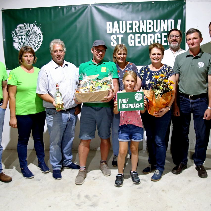 Eine Gruppe von Menschen in grüner Kleidung steht vor einem Banner mit der Aufschrift Bauernbund St. George. Ein Mann hält eine Flasche und einen Korb, während ein Mädchen ein Schild und Blumen hält.