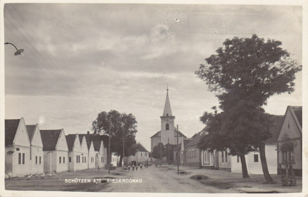 Ein altes Foto zeigt eine Dorfstraße mit einer Kirche, Bäumen und Gebäuden. Ein Schild zeigt 'Schiitzen A/G Niederdonau.' Menschen sind in der Ferne zu sehen.