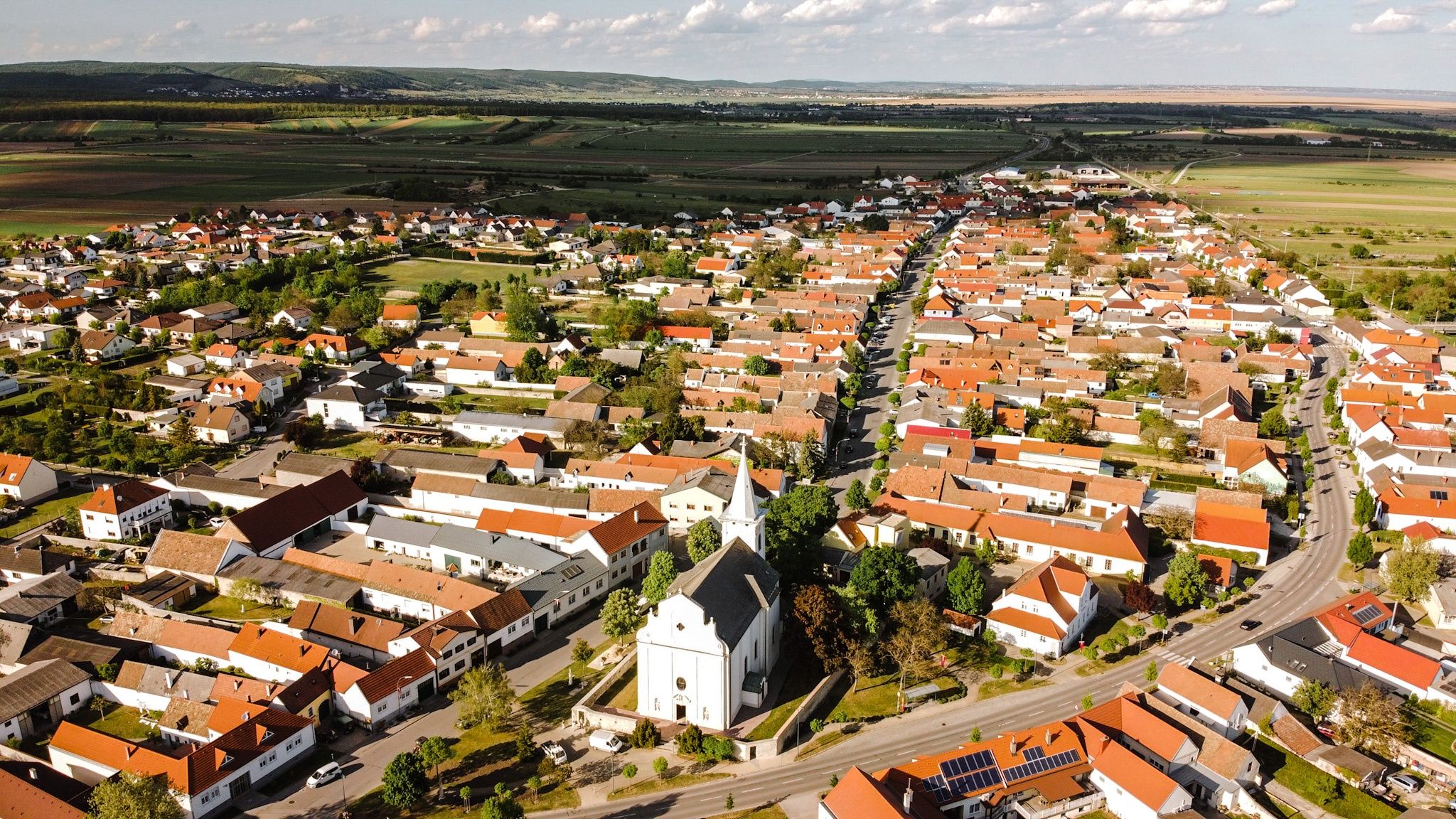 Luftaufnahme einer Stadt mit vielen Häusern, einer Kirche und einer Straße. Grüne Felder und Hügel umgeben das Gebiet.