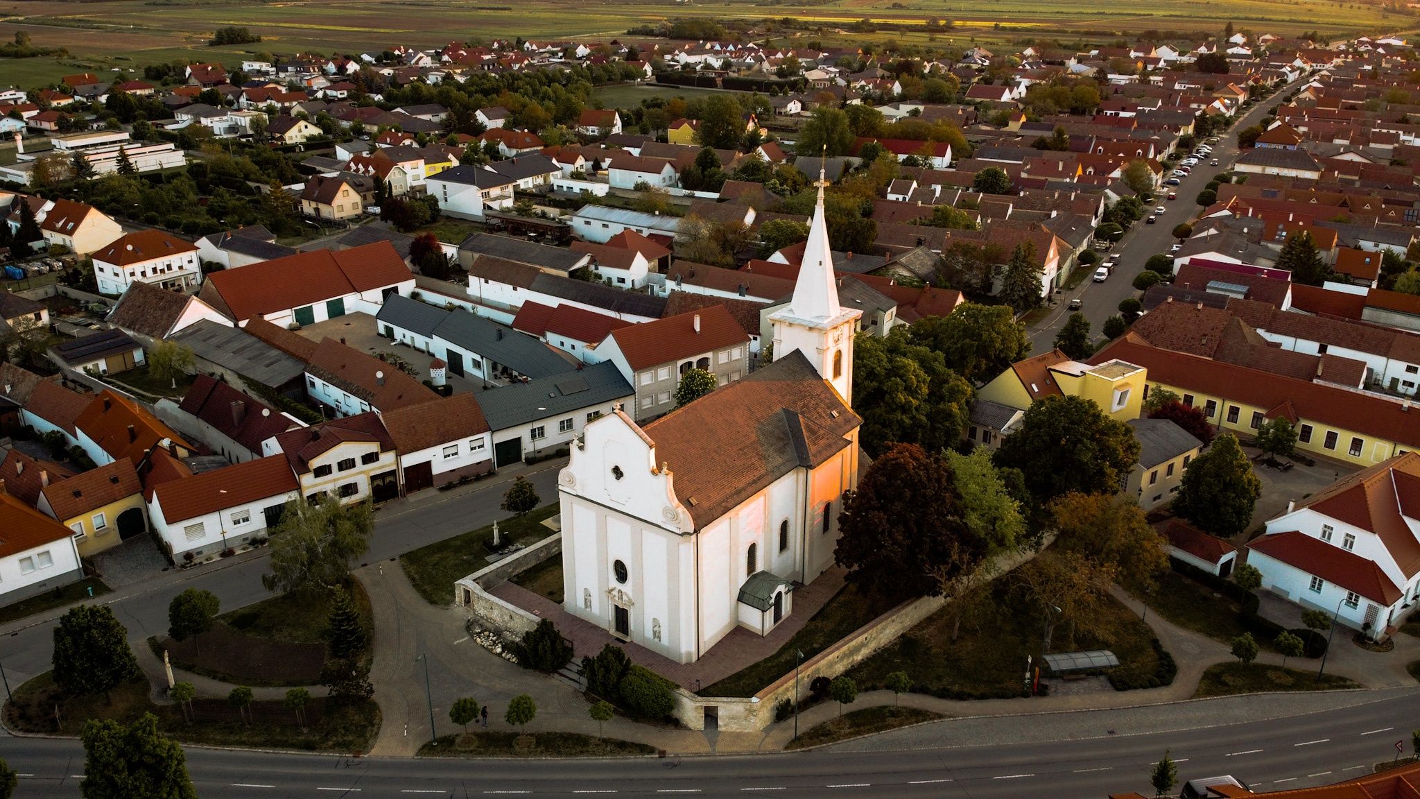 Luftaufnahme einer Stadt mit vielen Häusern und einer Kirche mit einem Kirchturm. Die Kirche ist von Bäumen und Büschen umgeben. Die Stadt hat viele Häuser und Gebäude mit unterschiedlich gefärbten Dächern. Die Straße ist im Vordergrund.