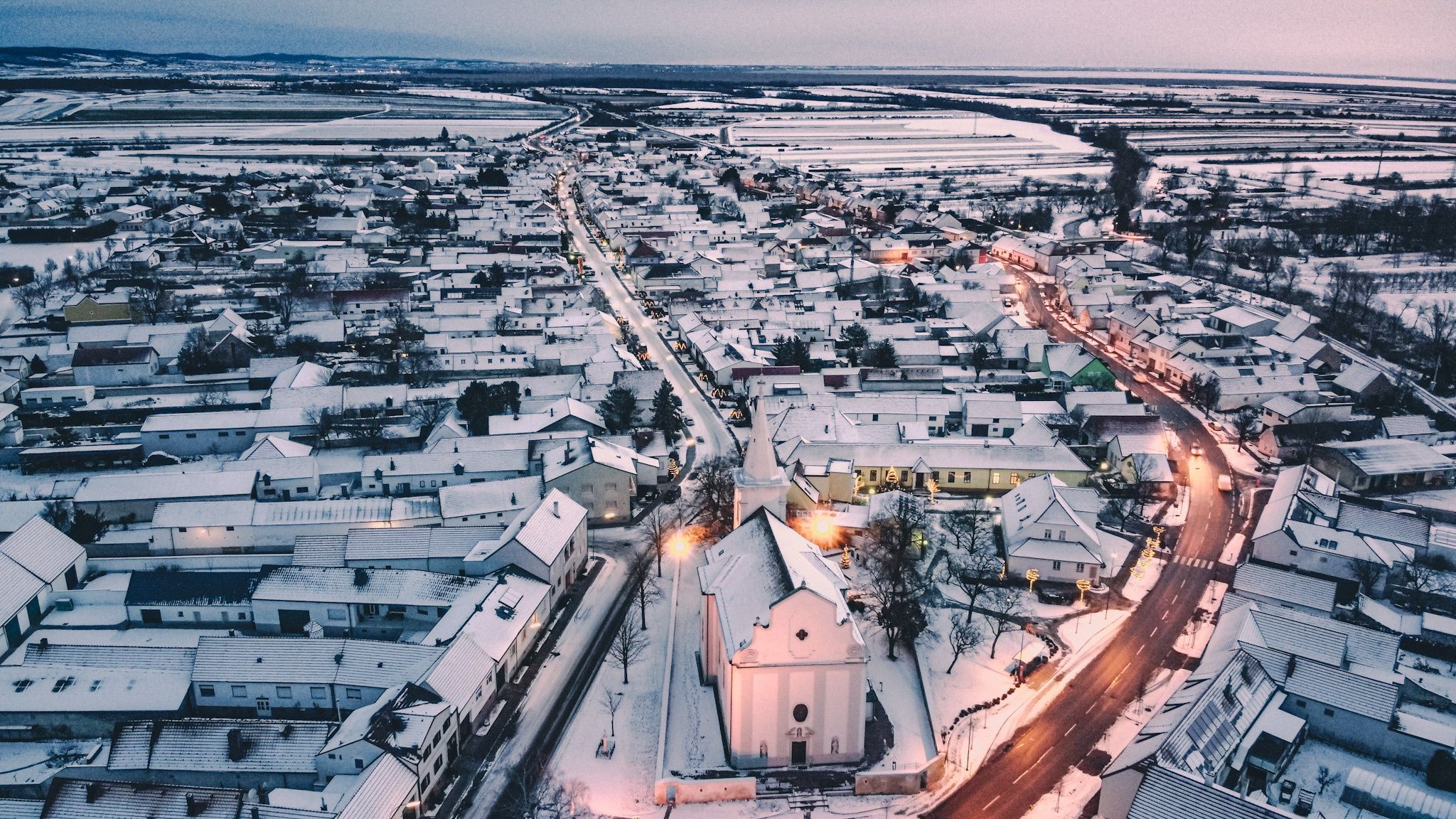 Eine Luftaufnahme einer Stadt mit einer schneebedeckten Kirche. Gebäude sind dicht gepackt, mit Straßen und Wegen, die mit Schnee bedeckt sind. Bäume umgeben die Kirche, und einige haben Schnee darauf.