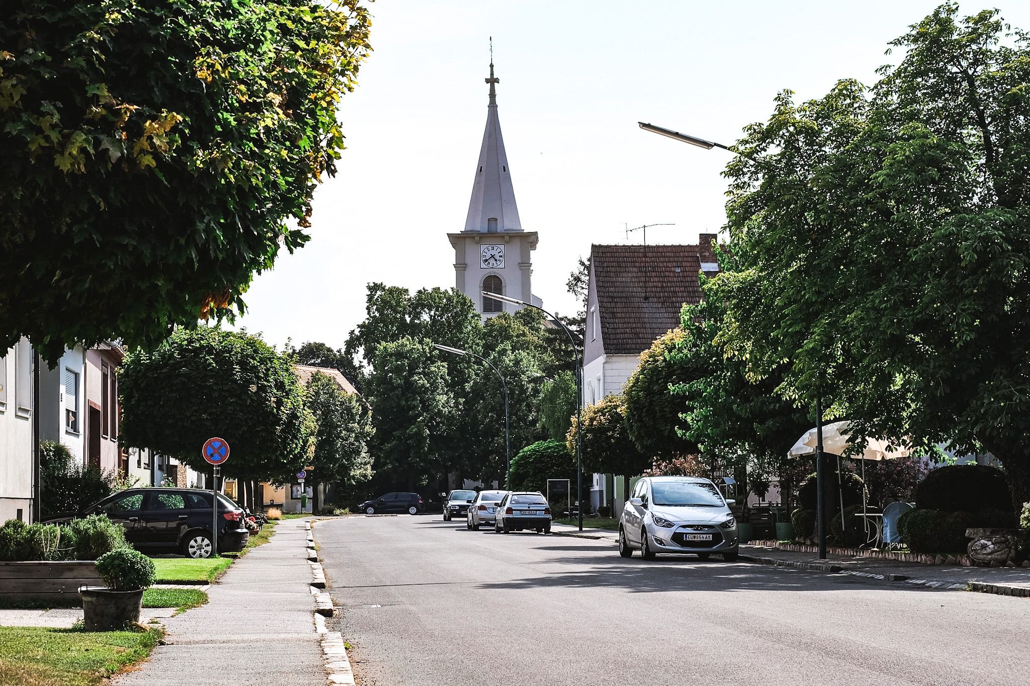 Eine Straße mit geparkten Autos und Bäumen auf beiden Seiten. Eine Kirche mit einem Turm und einer Uhr steht hinter den Autos.