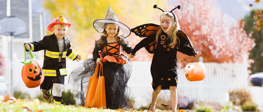 Drei Kinder spielen in einem Garten. Ein Mädchen hält eine Kürbis und ein anderes Mädchen einen Süßigkeitenbeutel. Der Junge trägt einen Feuerwehrmann-Kostüm.