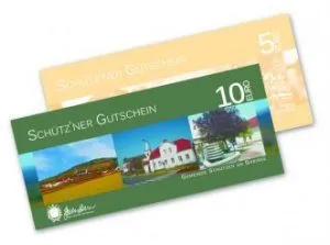 Eine grüne Schutzner Gutscheinkarte mit einem ländlichen Bild mit einem Haus und einem Baum. Sie trägt den Text 'SCHUTZNER GUTSCHEIN' und '10 EURO' auf der Vorderseite. Eine gelbe Schutzner Gutscheinkarte ist teilweise sichtbar.