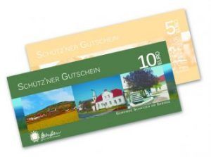 Eine grüne Schutzner Gutscheinkarte mit einem ländlichen Bild mit einem Haus und einem Baum. Sie trägt den Text 'SCHUTZNER GUTSCHEIN' und '10 EURO' auf der Vorderseite. Eine gelbe Schutzner Gutscheinkarte ist teilweise sichtbar.