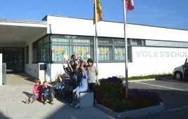 Kinder und Erwachsene sitzen auf einer Bank vor einem weißen Gebäude, einige winken. Hinter ihnen steht ein Fahnenmast mit einer Flagge. Das Gebäude steht 'VOLKSSCHUL'.