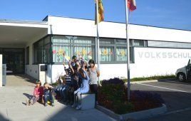 Kinder und Erwachsene sitzen auf einer Bank vor einem weißen Gebäude, einige winken. Hinter ihnen steht ein Fahnenmast mit einer Flagge. Das Gebäude steht 'VOLKSSCHUL'.