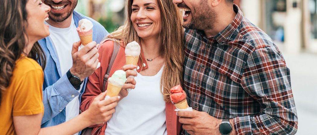 Eine Gruppe von Freunden steht draußen, lächelt und hält Eisbecher in den Händen. Die Frau in der Mitte hält zwei Eisbecher.