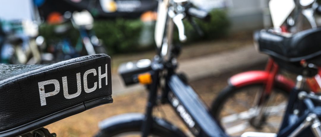 Ein Nahaufnahme eines schwarzen Rollers mit dem Wort 'PUCH' auf seinem Lenker, geparkt in einem Außenbereich mit einem unscharfen Hintergrund eines anderen Rollers.