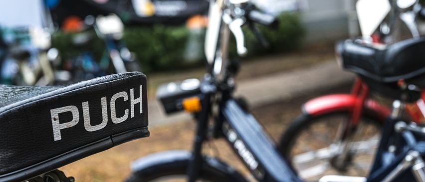 Ein Nahaufnahme eines schwarzen Rollers mit dem Wort 'PUCH' auf seinem Lenker, geparkt in einem Außenbereich mit einem unscharfen Hintergrund eines anderen Rollers.