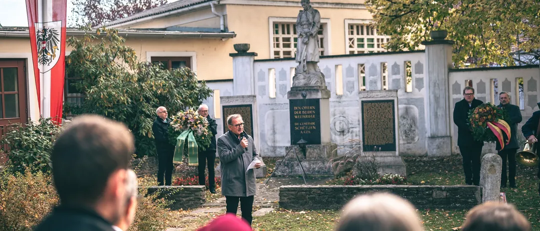 Vor einem Denkmal mit einer Statue und Tafeln findet eine öffentliche Veranstaltung statt, bei der ein Mann mit einem Mikrofon spricht, während andere zuhören.