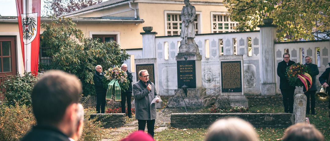 Vor einem Denkmal mit einer Statue und Tafeln findet eine öffentliche Veranstaltung statt, bei der ein Mann mit einem Mikrofon spricht, während andere zuhören.