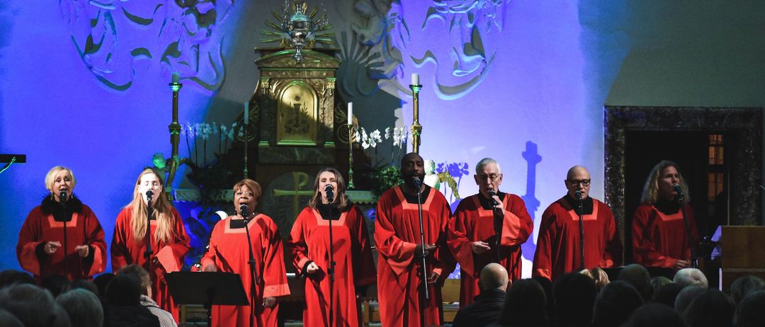 Ein Chor in roten Gewändern steht auf der Bühne und singt in Mikrofone. Dahinter ist ein goldener Altar von Kerzen beleuchtet, und dekorative Elemente schmücken die Wände. Das Publikum sitzt auf Kirchenbänken und beobachtet.