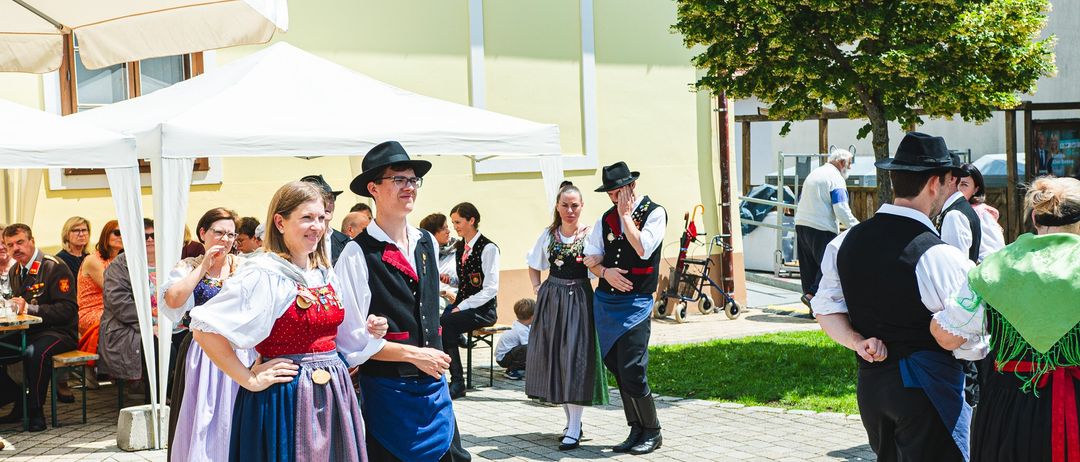 Mehrere Menschen in traditionellen Kostümen stehen vor einem Gebäude, einige unterhalten sich, während ein Kinderwagen in der Nähe steht. Die Sonne scheint hell und wirft Schatten auf den Boden.