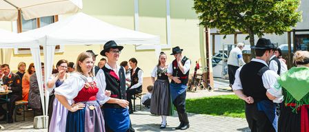 Mehrere Menschen in traditionellen Kostümen stehen vor einem Gebäude, einige unterhalten sich, während ein Kinderwagen in der Nähe steht. Die Sonne scheint hell und wirft Schatten auf den Boden.