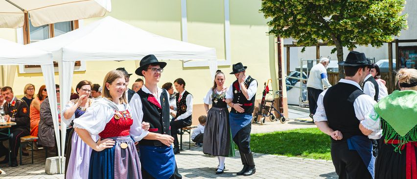 Mehrere Menschen in traditionellen Kostümen stehen vor einem Gebäude, einige unterhalten sich, während ein Kinderwagen in der Nähe steht. Die Sonne scheint hell und wirft Schatten auf den Boden.