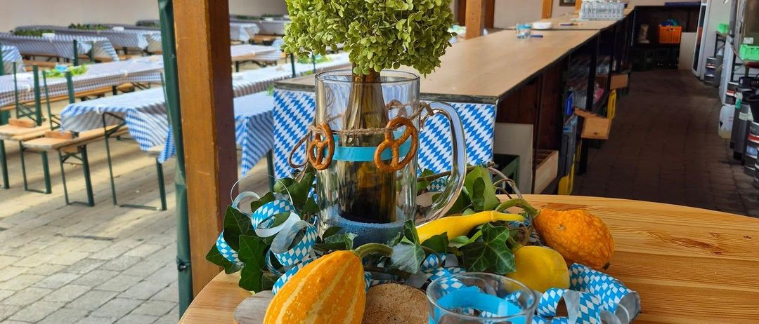 Ein hölzerner Tisch mit einem Krug, Brezeln, einer Kürbis und blauen Bändern. Dahinter ist ein Garten mit einem Ziegelboden und eine Wand mit grünen Bannern.