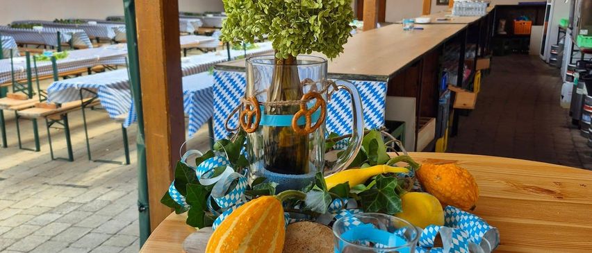 Ein hölzerner Tisch mit einem Krug, Brezeln, einer Kürbis und blauen Bändern. Dahinter ist ein Garten mit einem Ziegelboden und eine Wand mit grünen Bannern.