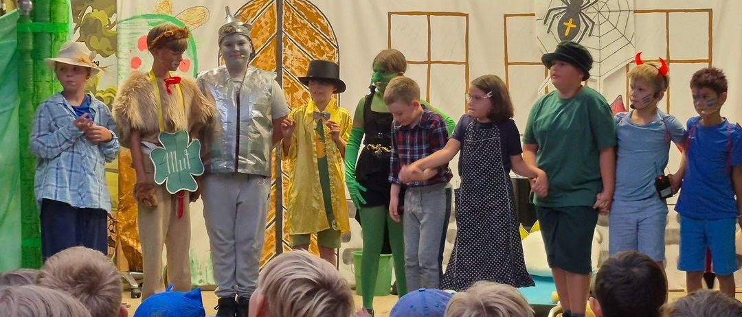 Eine Gruppe von Kindern in Kostümen führt ein Theaterstück vor einem Publikum auf, mit einem dekorierten Hintergrund und festlichen Lichtern darüber.