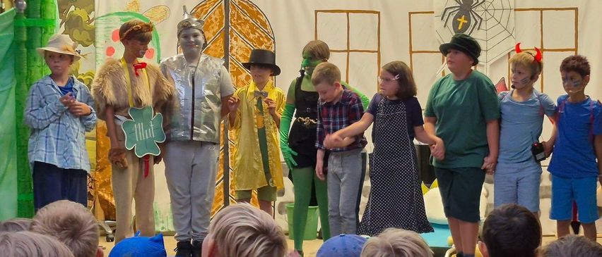 Eine Gruppe von Kindern in Kostümen führt ein Theaterstück vor einem Publikum auf, mit einem dekorierten Hintergrund und festlichen Lichtern darüber.