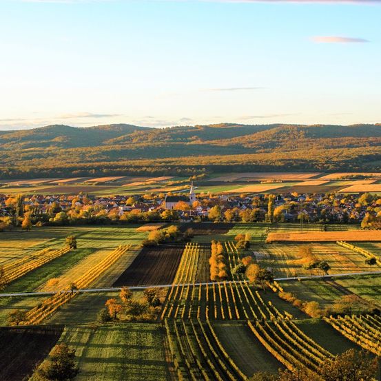 Luftaufnahme einer ländlichen Landschaft mit Feldern, einem Dorf und Hügeln unter klarem Himmel.