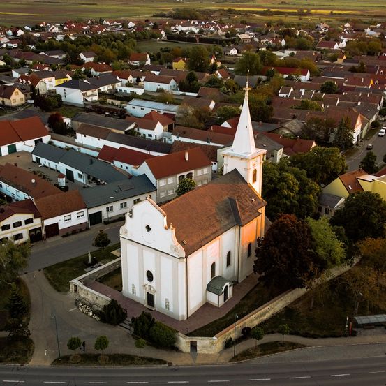 Luftaufnahme eines europäischen Dorfes mit einer Kirche im Zentrum, umgeben von Häusern mit roten Dächern und üppiger Grünfläche. Das Dorf liegt in einem Tal mit Feldern im Hintergrund.