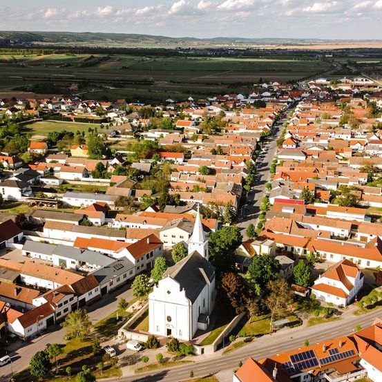 Luftaufnahme eines Dorfes mit vielen Häusern, einer Kirche und grünen Feldern. Die Häuser haben rote Dächer und sind von Bäumen umgeben. Die Kirche hat einen Turm.