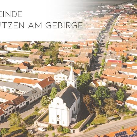 Eine Luftaufnahme einer Stadt mit vielen Häusern, die eine weiße Kirche mit einem Turm und roten Dächern zeigt. Die Stadt ist von Bäumen und Grünflächen umgeben, mit einer weißen Textüberlagerung oben 'Meinde'.