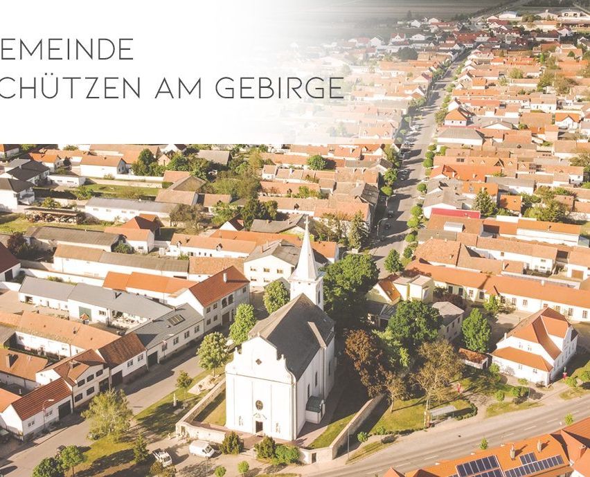 Eine Luftaufnahme einer Stadt mit vielen Häusern, die eine weiße Kirche mit einem Turm und roten Dächern zeigt. Die Stadt ist von Bäumen und Grünflächen umgeben, mit einer weißen Textüberlagerung oben 'Meinde'.