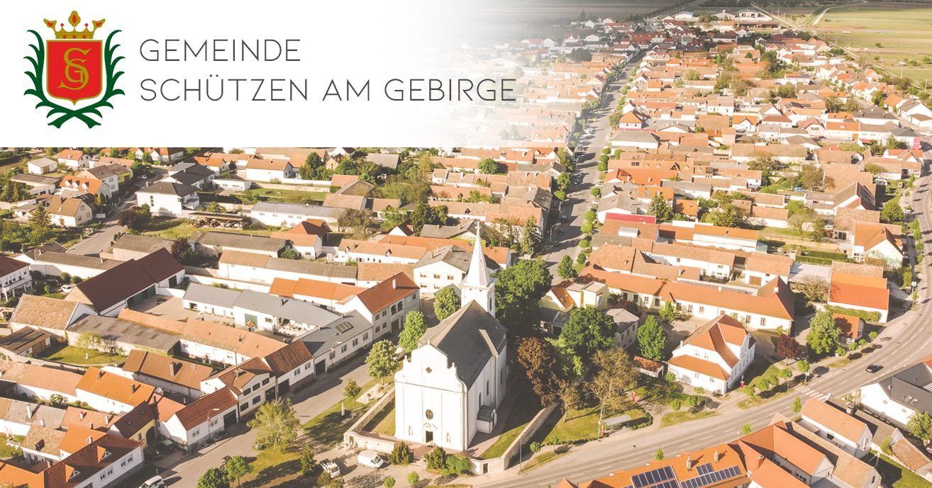 Eine Luftaufnahme einer Stadt mit vielen Häusern, die eine weiße Kirche mit einem Turm und roten Dächern zeigt. Die Stadt ist von Bäumen und Grünflächen umgeben, mit einer weißen Textüberlagerung oben 'Meinde'.