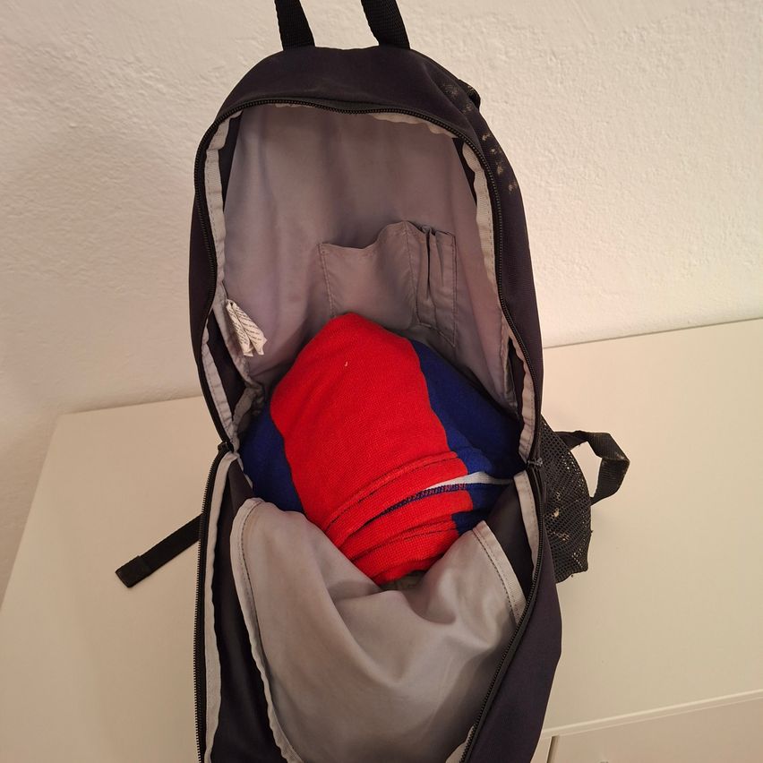 Ein offener Rucksack auf einer weißen Oberfläche zeigt ein rotes und blaues Tuch im Inneren. Der Rucksack ist dunkelblau mit einem grauen Fach und Reißverschlüssen.